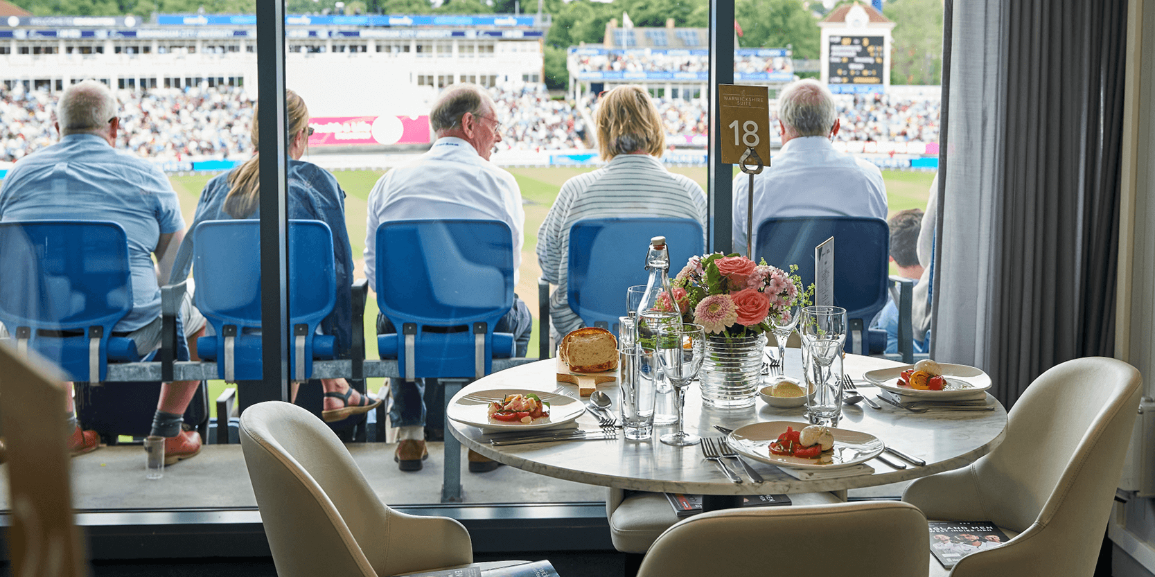View of a table set up in the Warwickshire Suite hospitality facility ay Edgbaston Stadium with a view of guests sat at their match seats overlooking the cricket pitch